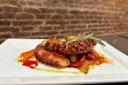 A dish featuring grilled octopus tentacles garnished with a sprig of rosemary, surrounded by roasted vegetables, including red peppers, on a white square plate. The background shows a brick wall.