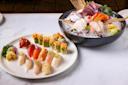 A platter of assorted sushi and sashimi on a marble surface. The sushi includes various types of nigiri with toppings like salmon and jalapeño. The sashimi bowl features sliced fish on ice with garnishes and wasabi.