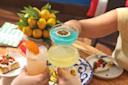 Three people toast with colorful cocktails—one blue drink with a dried lemon slice, one yellow drink, and one orange drink—over a table set with appetizers, a plate of lemons, and a green striped book.