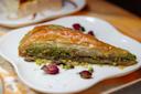 A close-up of a slice of baklava on a white plate, topped and filled with crushed pistachios, with dried rosebuds and pistachio crumbs scattered around as garnish.