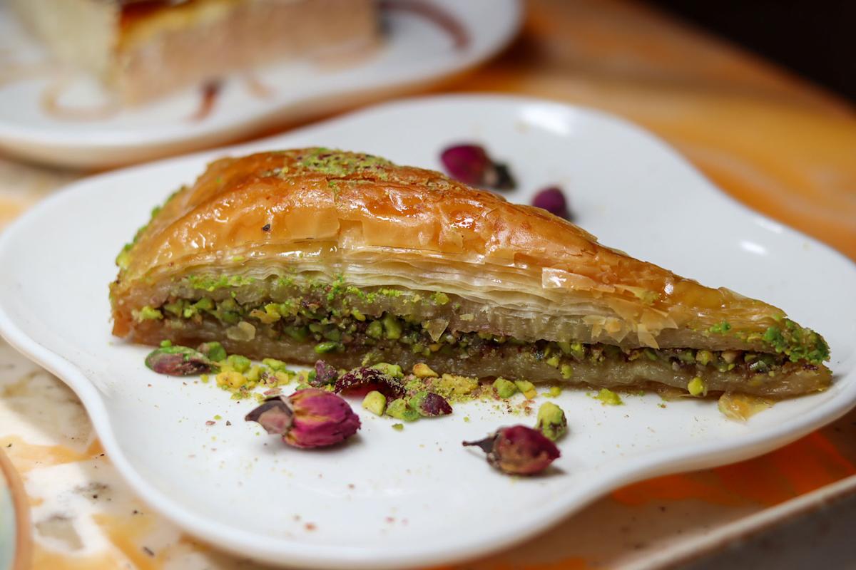 A close-up of a slice of baklava on a white plate, topped and filled with crushed pistachios, with dried rosebuds and pistachio crumbs scattered around as garnish.