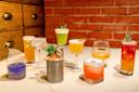 A variety of colorful cocktails in different glasses are displayed on a white surface in front of a red brick wall, each garnished uniquely with fruit, herbs, or leaves.