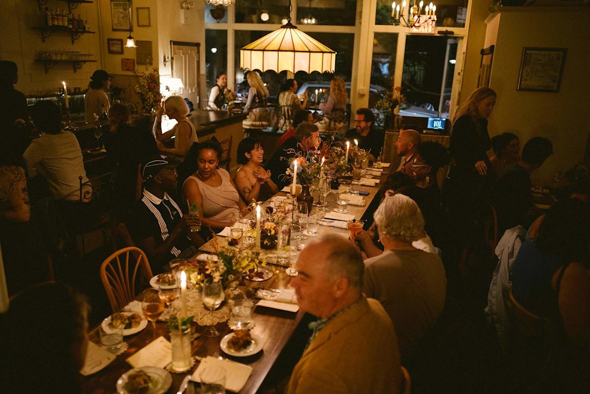 A large group of people sit at long, candle-lit tables in a cozy, warmly lit restaurant, enjoying food, drinks, and conversation in a lively, intimate atmosphere.