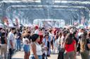 A large crowd of people gathers under a covered pavilion at the The Original Latin Food Festival with colorful vendor tents, flags, and a stage in the background. The atmosphere is lively and festive.
