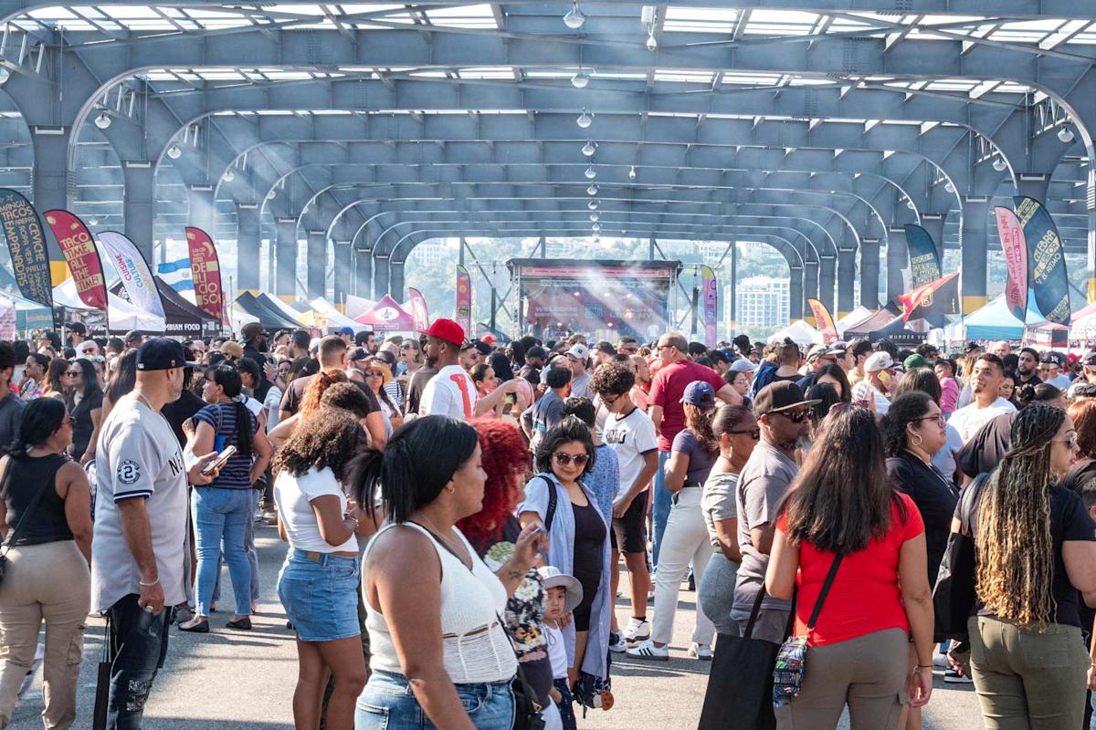 A large crowd of people gathers under a covered pavilion at the The Original Latin Food Festival with colorful vendor tents, flags, and a stage in the background. The atmosphere is lively and festive.