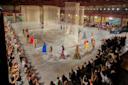 A fashion show features models walking a circular runway. The models wear various colorful outfits against a backdrop of vertical light-colored string installations. The audience surrounds the runway, capturing photos.