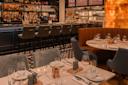 Cozy restaurant interior with elegantly set tables, cushioned chairs, and a bar lined with high-backed stools. Warm lighting highlights the decor, featuring neatly arranged glassware and tableware. Shelves behind the bar display an assortment of bottles.