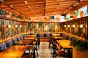A cozy restaurant interior with wooden tables and chairs, vibrant patterned walls, and warm hanging lights. The ceiling has exposed beams, and plants decorate the high shelves, complementing the artistic atmosphere.
