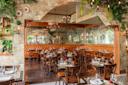 Elegant restaurant interior with rustic decor, featuring brown wooden tables and chairs, tan cushioned seating, and a mirrored wall. Greenery and chandeliers add to the cozy, sophisticated atmosphere. Tables are set with white plates and glassware.