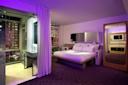 A modern hotel room with a large bed, minimalist furniture, purple accent lighting, a wall-mounted TV, and a glass-walled bathroom with a city view through floor-to-ceiling windows.