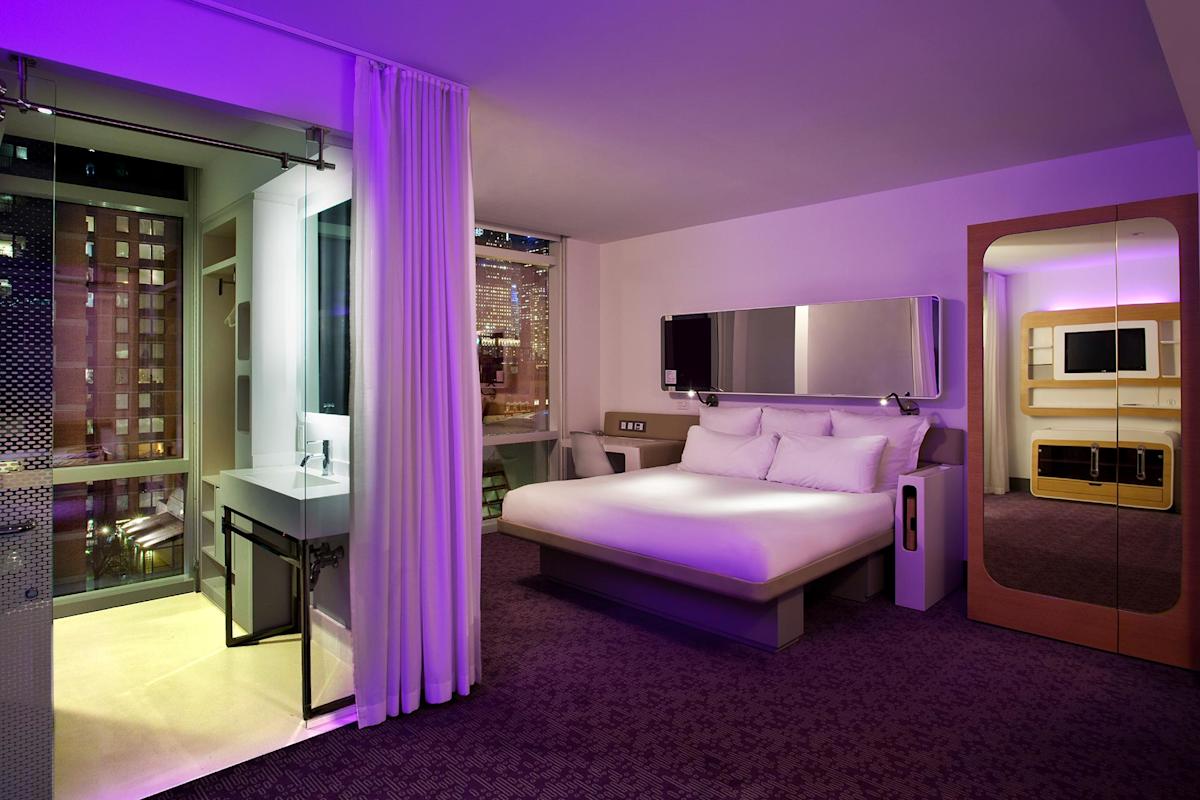 A modern hotel room with a large bed, minimalist furniture, purple accent lighting, a wall-mounted TV, and a glass-walled bathroom with a city view through floor-to-ceiling windows.