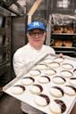 A baker wearing glasses, a blue cap, and a white coat holds a large tray of black-and-white cookies in a commercial kitchen, with racks of bread and ovens visible in the background.