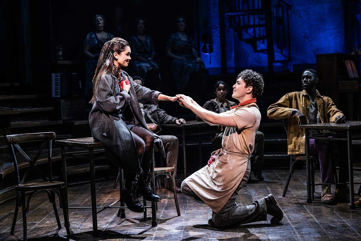 A man kneels and holds hands with a woman sitting on a table, both performing on a dimly lit theater stage with other cast members seated in the background.