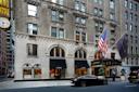 Street view of a historic hotel with tan brick facade, large arched windows, two flags (American and hotel), a valet standing by the entrance, and a black car parked in front.