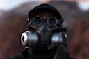 A person wearing dark round goggles, a black cap, and a black gas mask with large silver filters, dressed in black clothing and gloves, stands outdoors with a blurred rocky background.