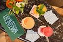 A marble table with three colorful cocktails, a stack of patterned coasters, a bouquet of flowers, and a green menu with "LEONESSA" written on it in white letters.