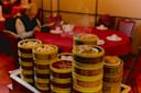 A stack of bamboo steamers filled with dim sum sits on a cart in a restaurant, with steam rising. In the background, a person sits at a red table reading a menu.