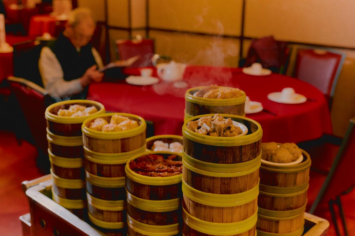 A stack of bamboo steamers filled with dim sum sits on a cart in a restaurant, with steam rising. In the background, a person sits at a red table reading a menu.