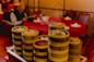 A stack of bamboo steamers filled with dim sum sits on a cart in a restaurant, with steam rising. In the background, a person sits at a red table reading a menu.