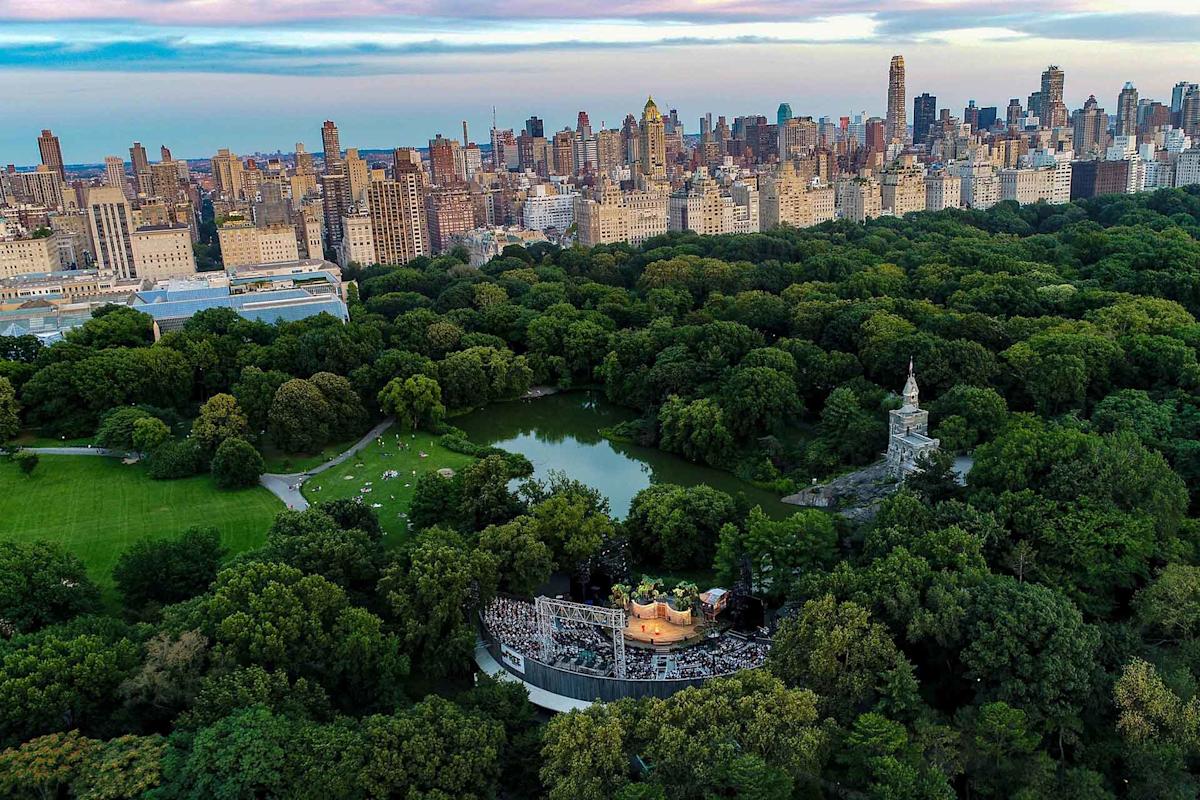 Aerial view of Central Park in New York City, showing a lush green landscape, a pond, an outdoor theater with an audience, and the city skyline in the background under a blue sky.