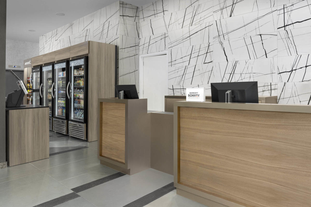 A modern hotel reception area with a wooden desk, computer monitors, and a small sign that reads "Bonvoy." On the left, there is a convenience section with refrigerated drinks and snacks in glass-door coolers