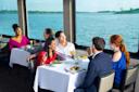 A group of six people dressed in semi-formal attire sit at tables on a boat, enjoying food and drinks while smiling and talking, with large windows showing a scenic view of a lake and trees.