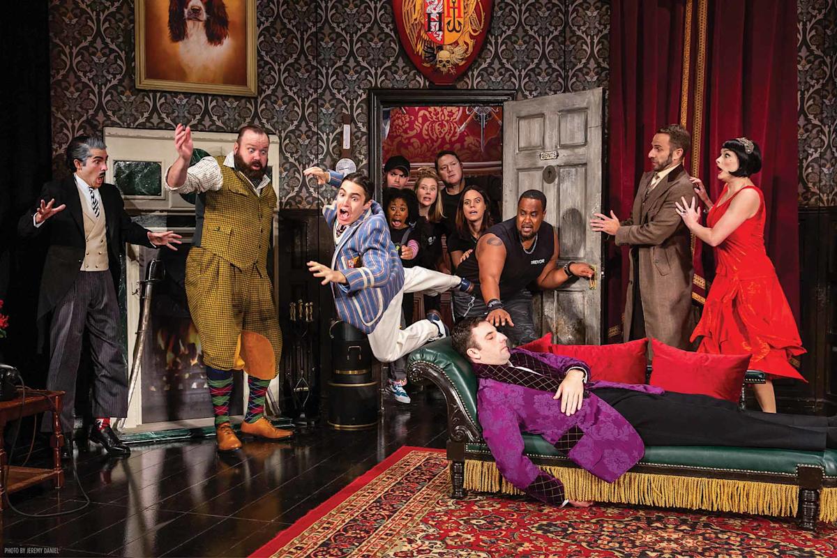 A chaotic stage scene: one man tumbles off a green couch as others react in shock; actors in period costumes fill the ornate room, with dramatic expressions and lively movement.