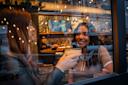 Two women sit at a bar table seen through a window, smiling and holding cocktails. Warm lights and bar shelves create a cozy atmosphere. Reflections of the street add depth to the scene.