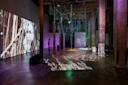An art installation in an industrial space features hanging green ropes, concrete pillars, and projected images of roots and branches on walls and floor, with a person observing the exhibit.