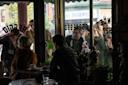 People gather outside a cafe window, pressing their faces against the glass, looking inside. Inside, two people sit at a table with cups and saucers.