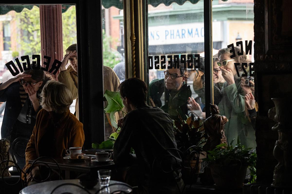 People gather outside a cafe window, pressing their faces against the glass, looking inside. Inside, two people sit at a table with cups and saucers.