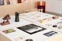 A display table features art books, prints, ceramics, sculptures, shells, and small architectural objects in a modern, minimalist gallery with light wood surfaces and a red velvet chair in the background.