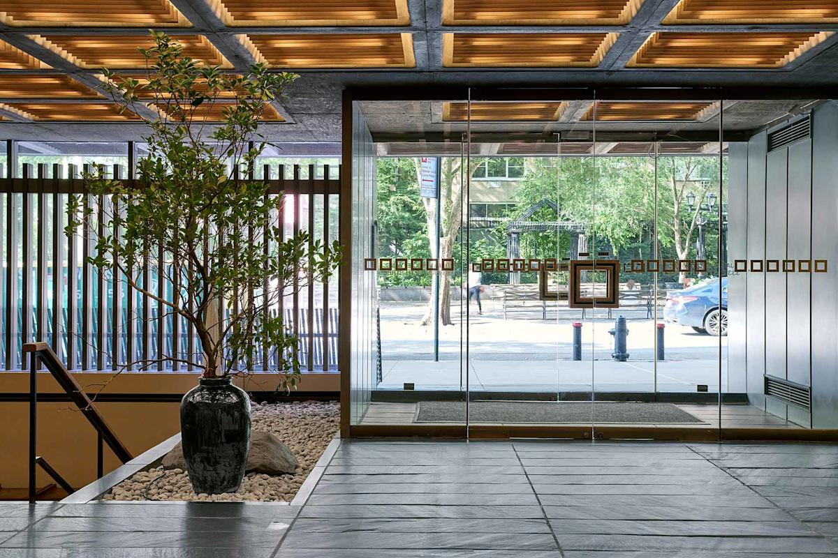 Modern building lobby with large glass doors, a potted tree on a bed of stones, and wood-paneled ceiling. Sunlight and greenery can be seen outside through the glass entrance.
