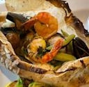 Seafood including shrimp, mussels, and clams, along with vegetables, served in parchment paper that is opened and slightly browned from baking.