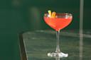 A bright pink cocktail in a coupe glass sits on a dark green marble surface, garnished with a twist of orange peel.