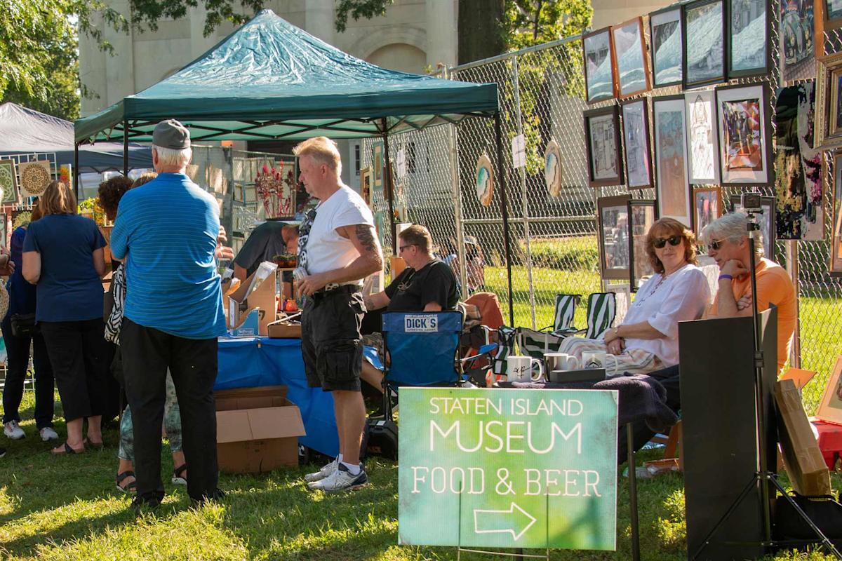 People gather near vendor tents at an outdoor fair. Art is displayed on fences and people sit near a sign that reads “Staten Island Museum Food & Beer.” The event takes place on a sunny day.