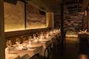 A dimly lit restaurant with exposed brick walls and elegant table settings. Each table has white tablecloths, wine glasses, and small flower arrangements. Black chairs line the tables, creating a cozy and intimate dining atmosphere.