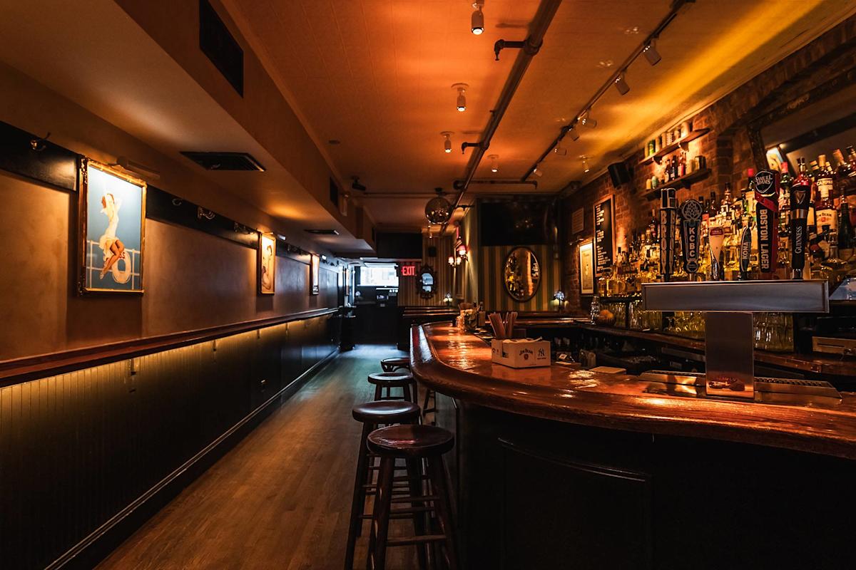 A dimly lit bar with wooden stools along a curved counter, liquor bottles, and warm ambient lighting. Framed art hangs on the left wall, and the bar appears empty and inviting.