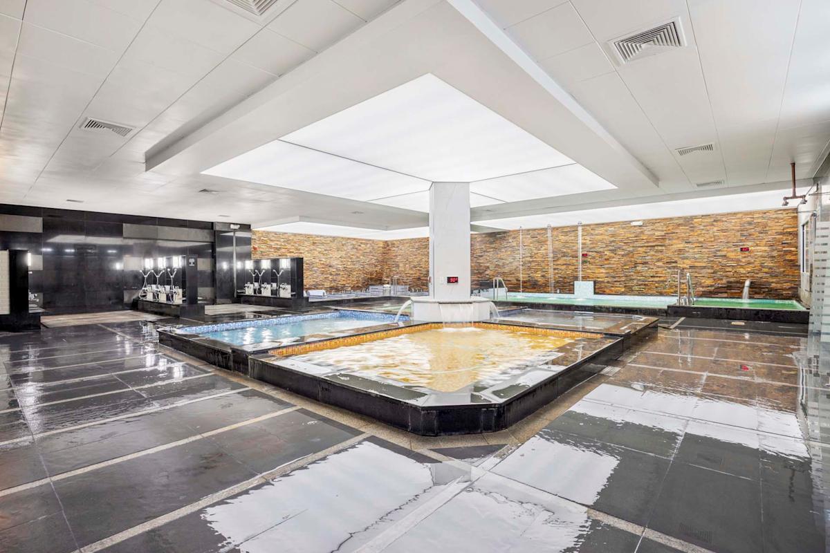 A spacious indoor spa with multiple rectangular pools, modern black and white decor, stone accent walls, and large ceiling lights reflecting on the wet tiled floor.