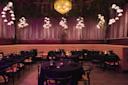 A dimly lit restaurant interior with round tables and chairs, adorned with dark tablecloths. Soft lighting from hanging floral-style lamps and a central chandelier create a cozy ambiance. Plush tufted seating lines the walls beneath curtain drapes.