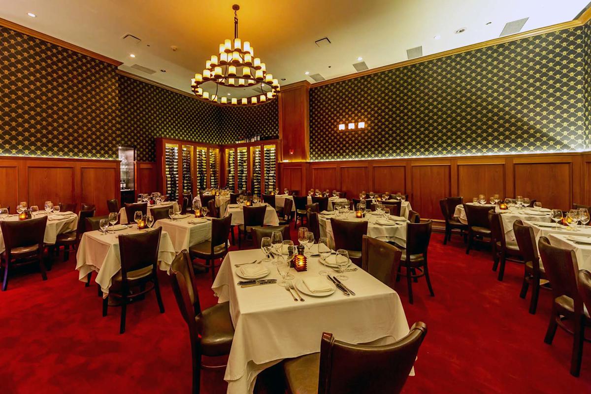 Elegant restaurant dining room with red carpet, dark wood paneling, tables covered with white cloths, neatly set with plates and glasses, wine racks along the back wall, and a large chandelier overhead.