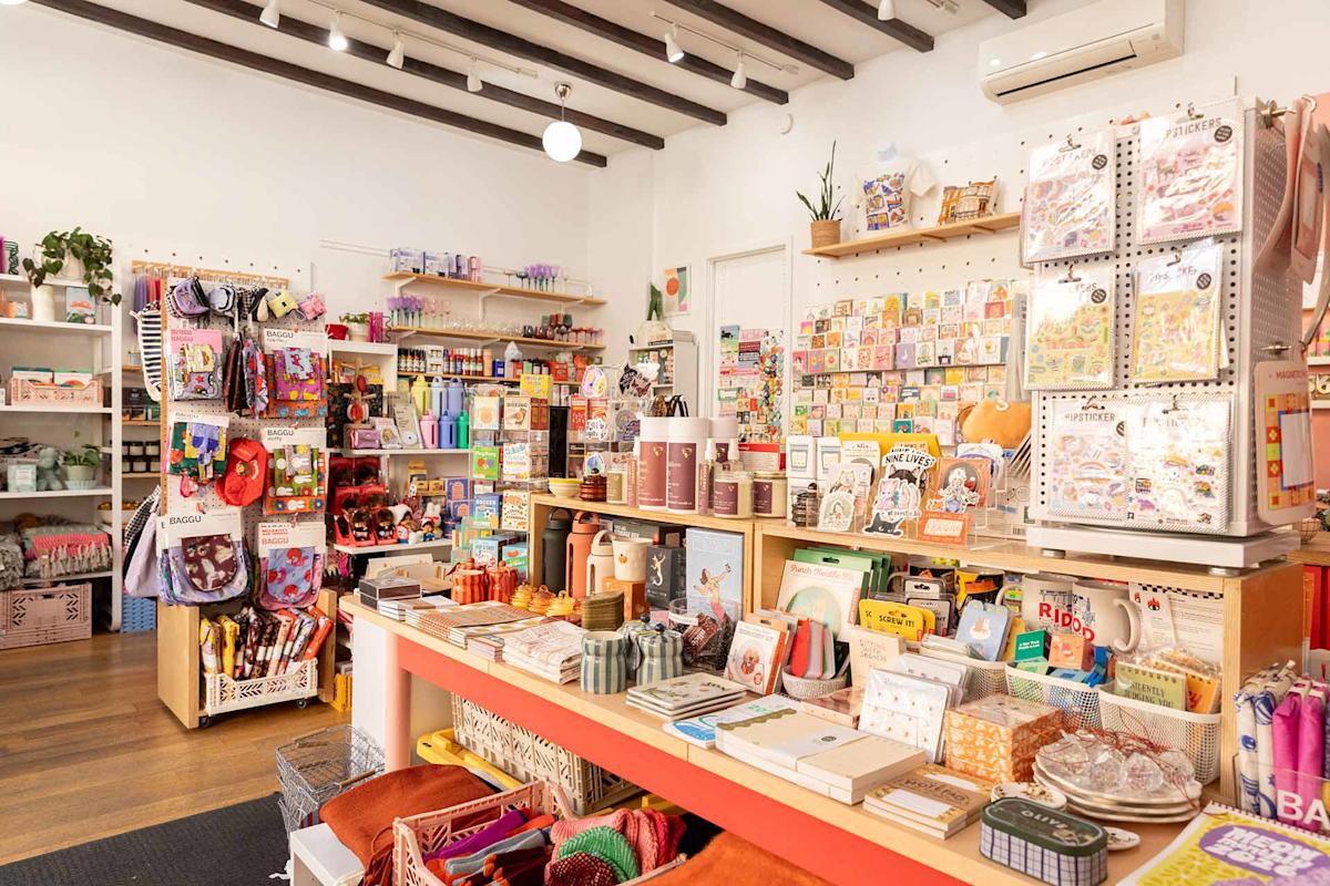 Bright stationery shop interior with colorful notebooks, pens, greeting cards, art supplies, and gift items displayed on shelves and tables, with wooden floors and white walls.