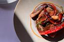 A plate with grilled red peppers, a slice of seared meat, and a clove of garlic, garnished with herbs and sauce, shown in bright natural light with shadows on a light-colored plate and table.