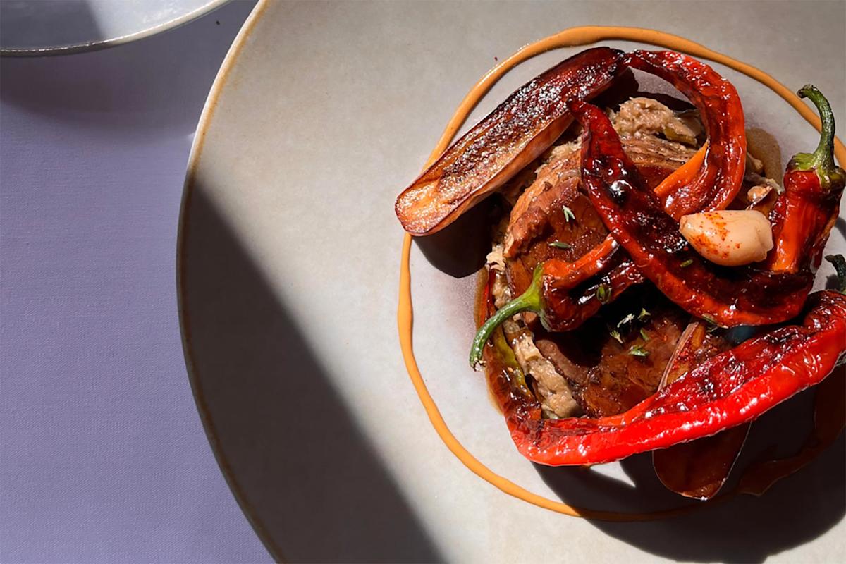 A plate with grilled red peppers, a slice of seared meat, and a clove of garlic, garnished with herbs and sauce, shown in bright natural light with shadows on a light-colored plate and table.