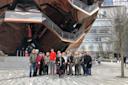A group of people posing for a photo in front of a modern, metallic structure with overlapping triangular panels. The building has staircases and reflective surfaces