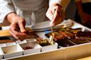A chef's hands preparing a dish, with one hand holding a spoon over a container with herbs. Grilled meat rests on a rack beside several small bowls of spices and herbs. Plates are stacked in the background.