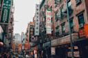 A busy street in Chinatown lined with restaurants and shops, featuring colorful signs in Chinese and English, fire escapes on brick buildings, and people walking on the sidewalks.