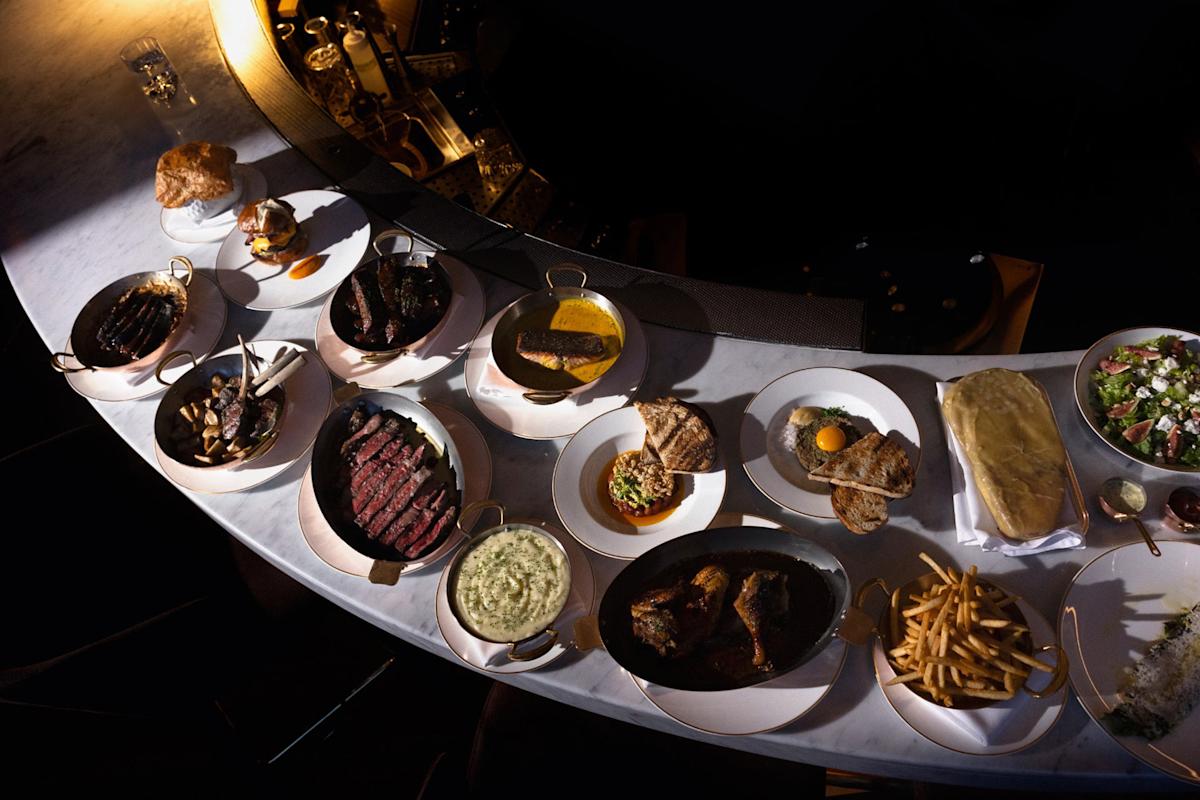 A curved marble table set with various dishes including steaks, grilled vegetables, fries, salads, bread, a burger, creamed spinach, and dessert, with dim lighting and elegant plating.