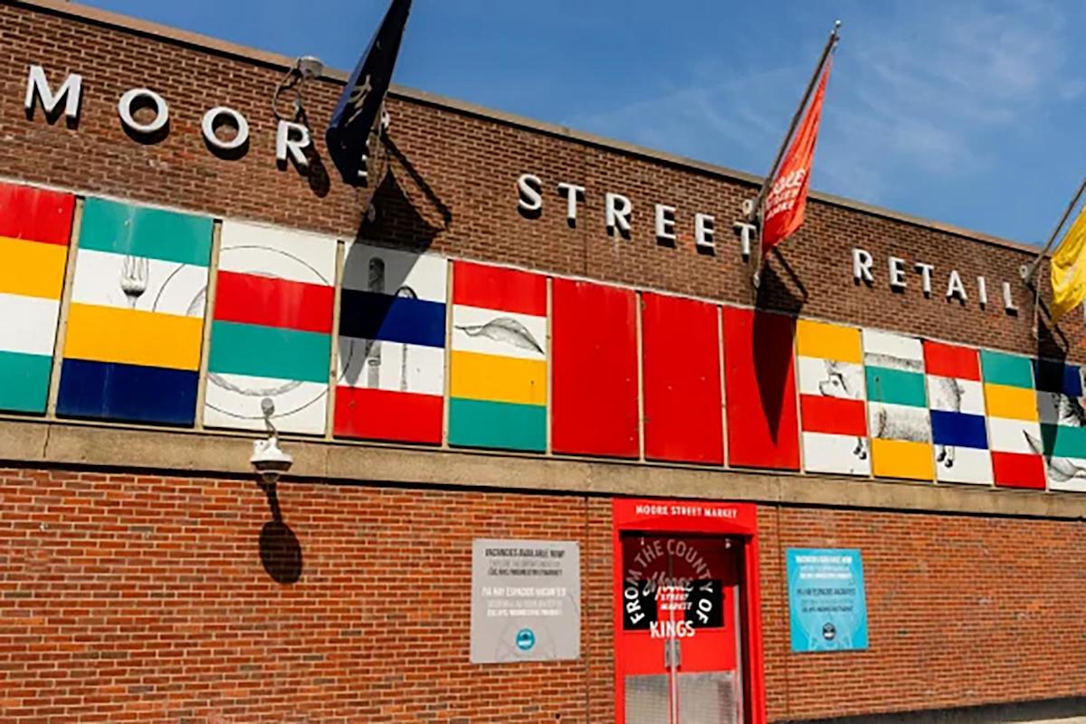 A brick building labeled "Moore Street Retail" features colorful rectangular panels, flags, and two informational signs near a red entrance door under a blue sky.