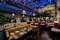 A stylish, dimly lit restaurant interior featuring wooden tables set with white plates and pink napkins. Brown leather booths and wooden chairs surround the tables. 